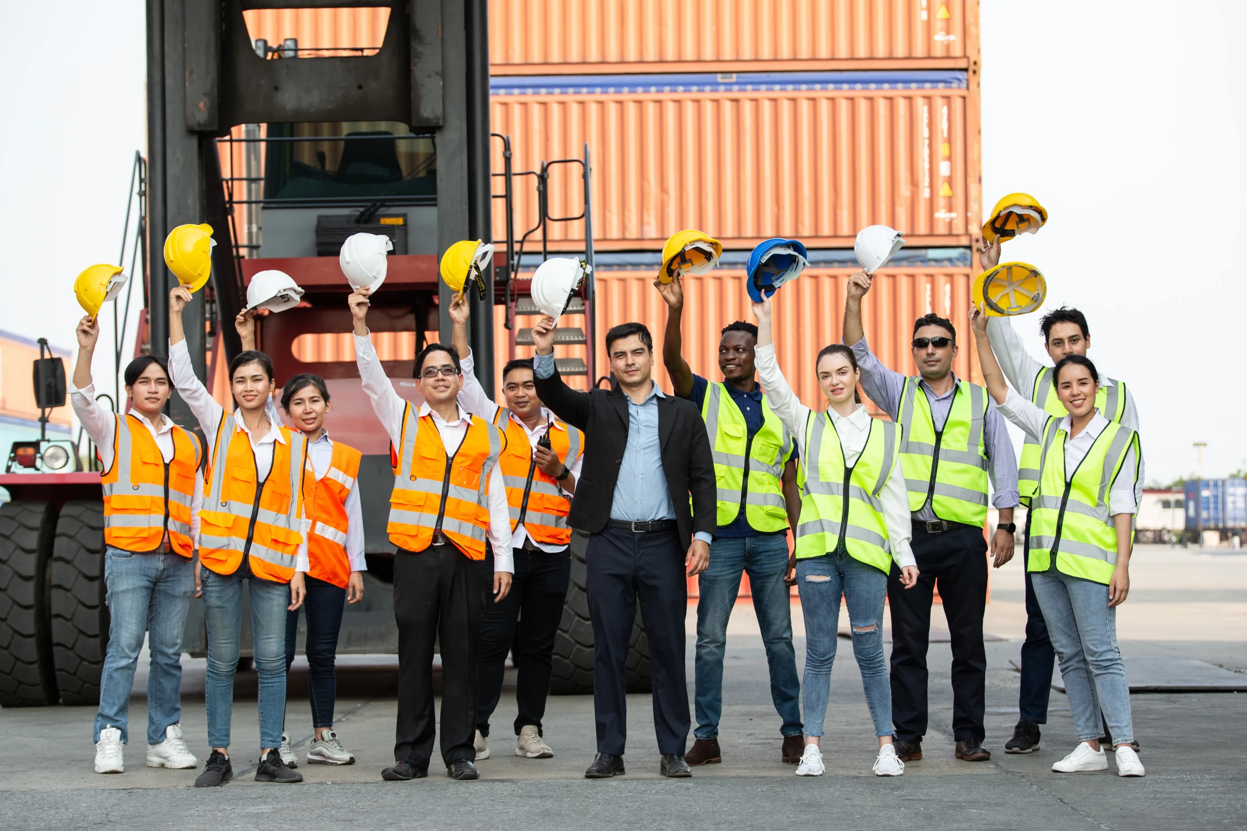 A diverse group of logistic workers in safety vests raising their hard hats in front of stacked shipping containers, showcasing teamwork.