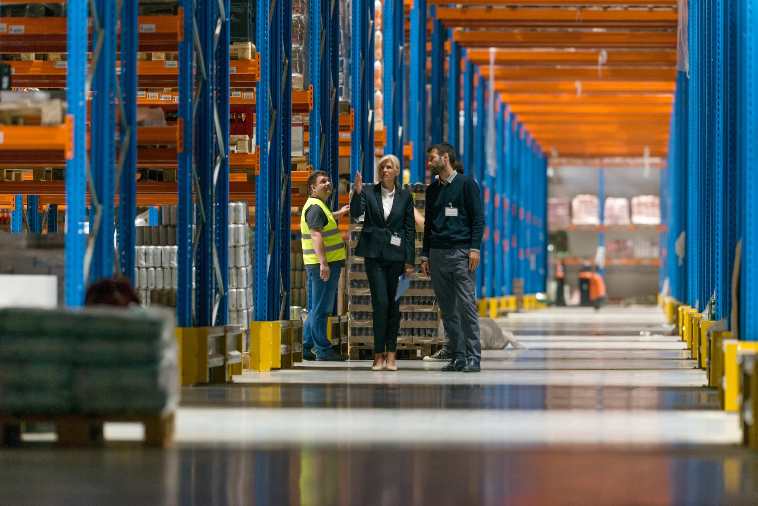 Team discussing inventory management in a spacious duty-free warehouse with tall blue and orange shelving.