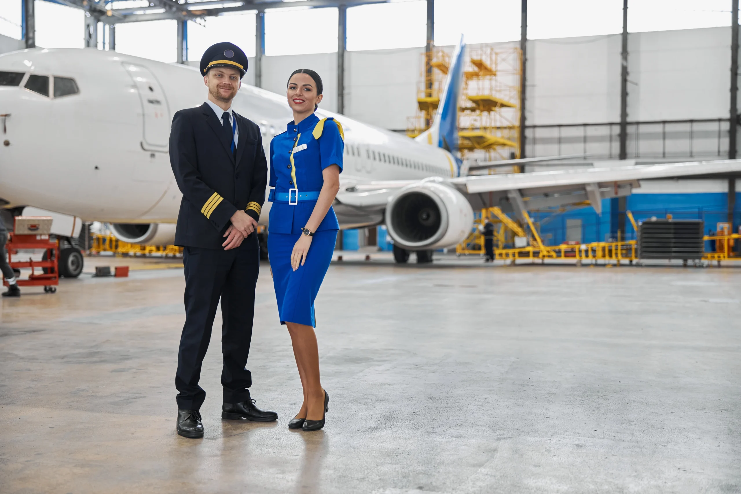 A pilot and cabin crew member standing in front of an airplane inside a hangar, representing the human element in airline maintenance and logistics operations.