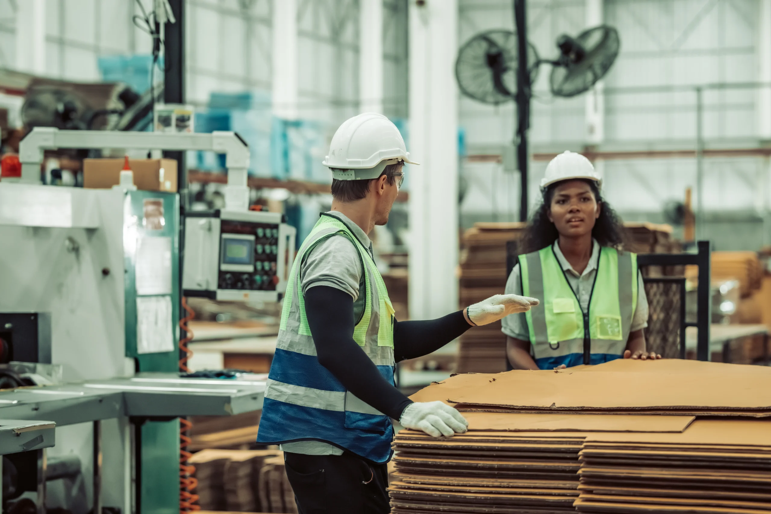 Factory workers discussing precision in cardboard preparation for packaging, aligning with compliance in regulated industries logistics.