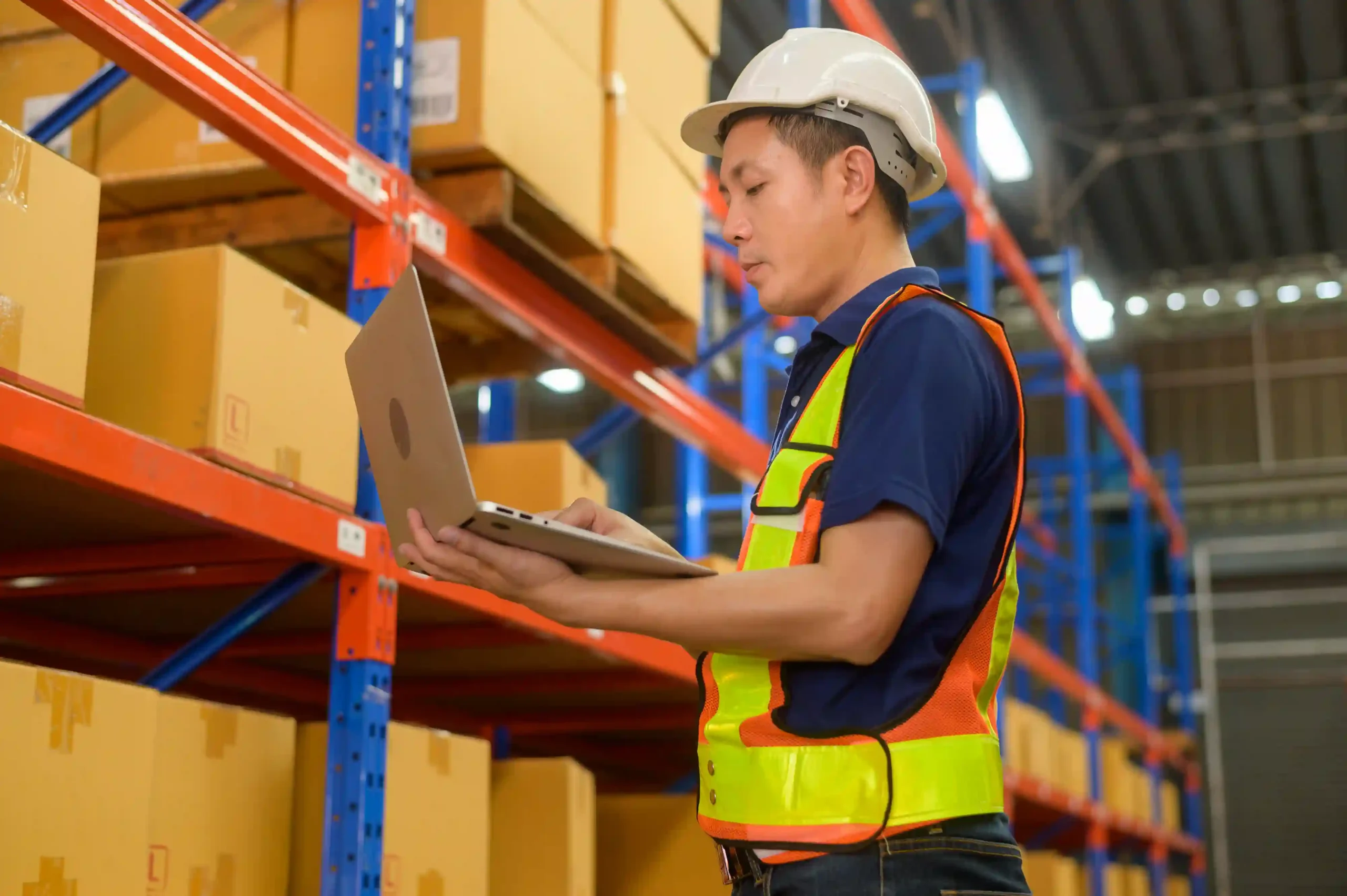 A warehouse worker wearing a safety vest and helmet uses a laptop to check inventory. This image represents real-time data tracking and efficiency with a warehouse management system questionnaire.