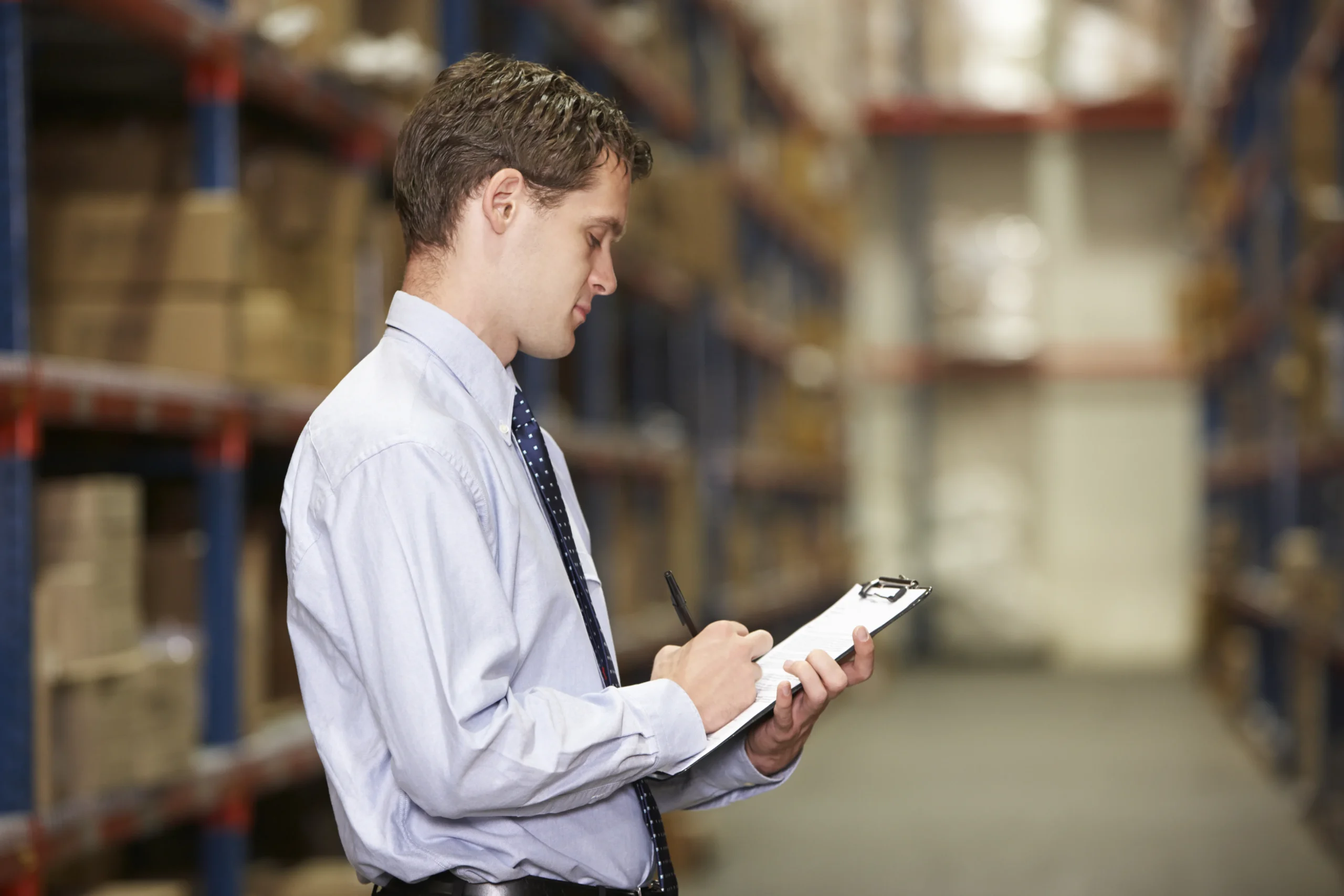 A warehouse manager in a business suit reviewing an inventory management system journal on a clipboard, ensuring stock accuracy and efficient logistics.