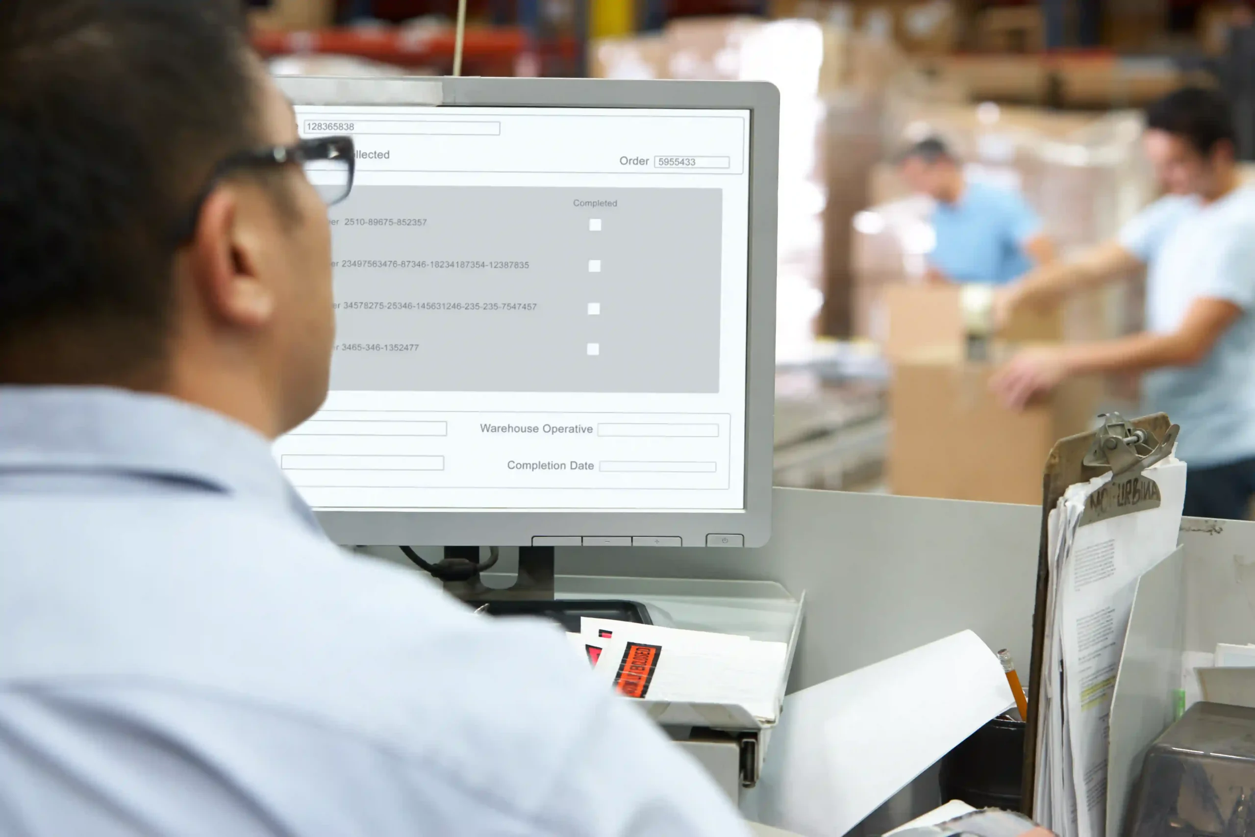 A logistics professional monitors order tracking on a desktop screen in a busy warehouse, illustrating the importance of a warehouse management system questionnaire in optimizing operations.