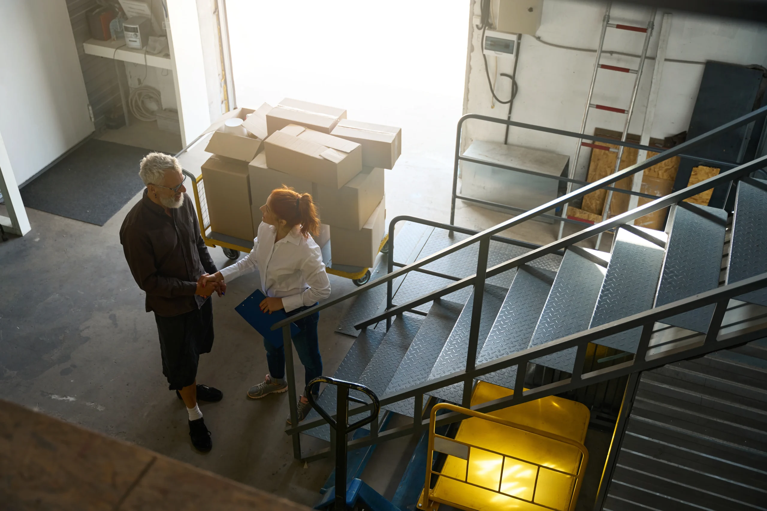 Red-haired logistics manager with a blue folder shaking hands with a delivery driver near shipment boxes.