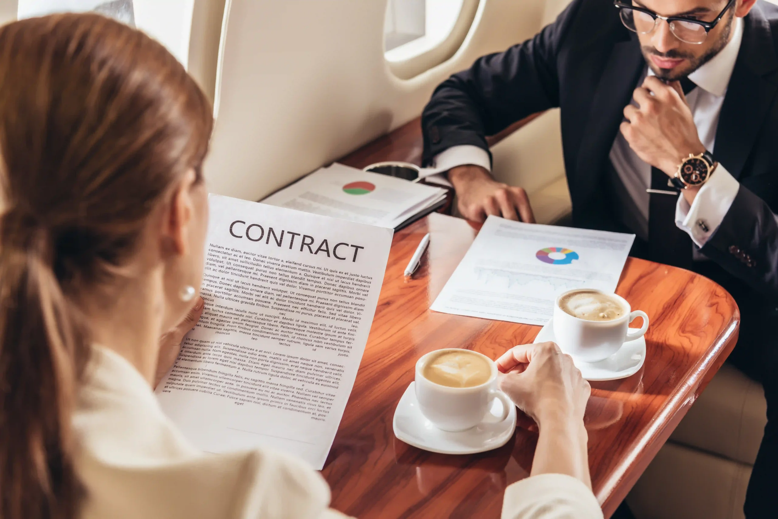 A businesswoman and a businessman sit inside a private meeting space, reviewing and discussing the details of a logistics contract agreement over coffee.
