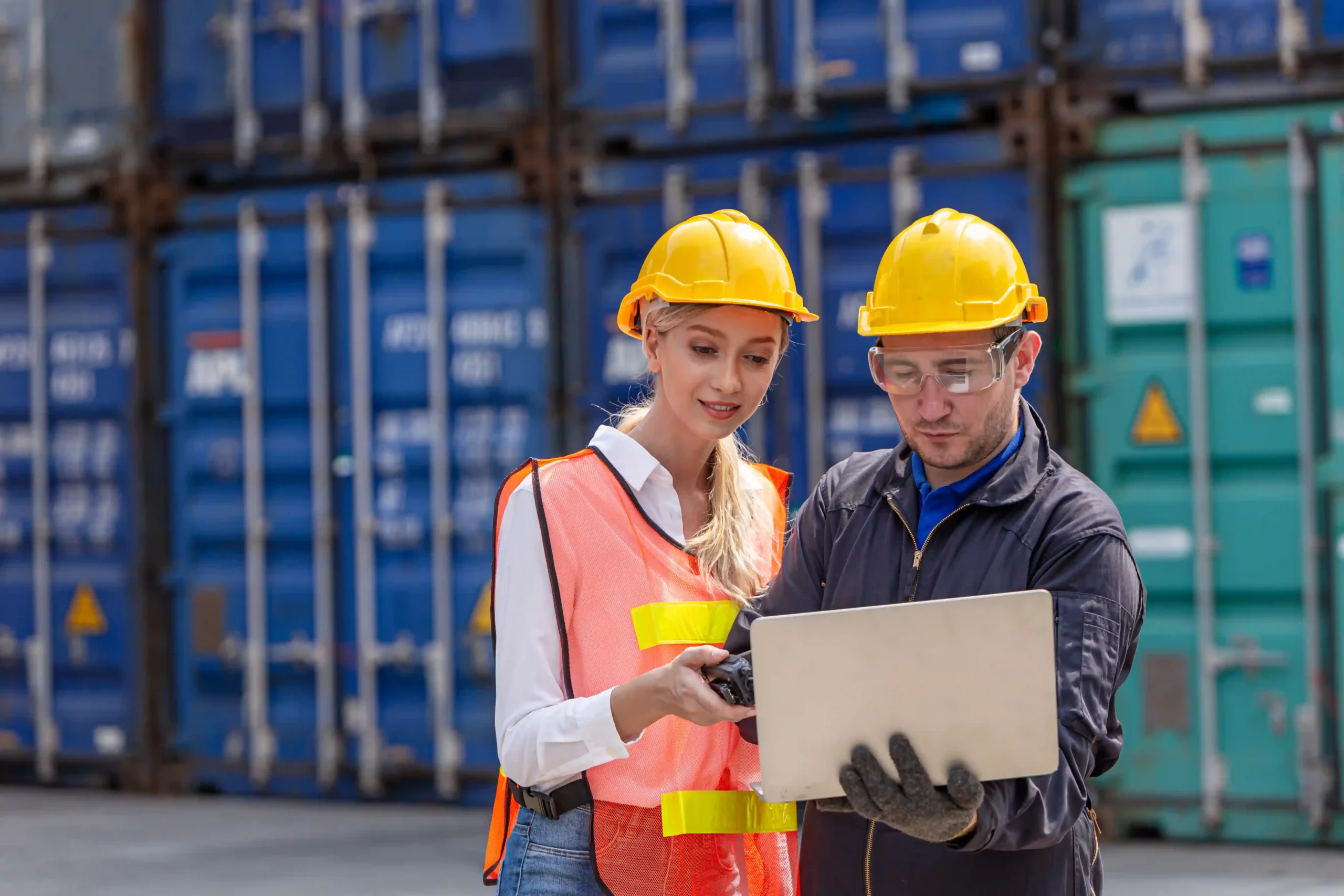 Engineers analyzing data on a laptop at a cargo shipping yard, utilizing custom report design for supply chain management.