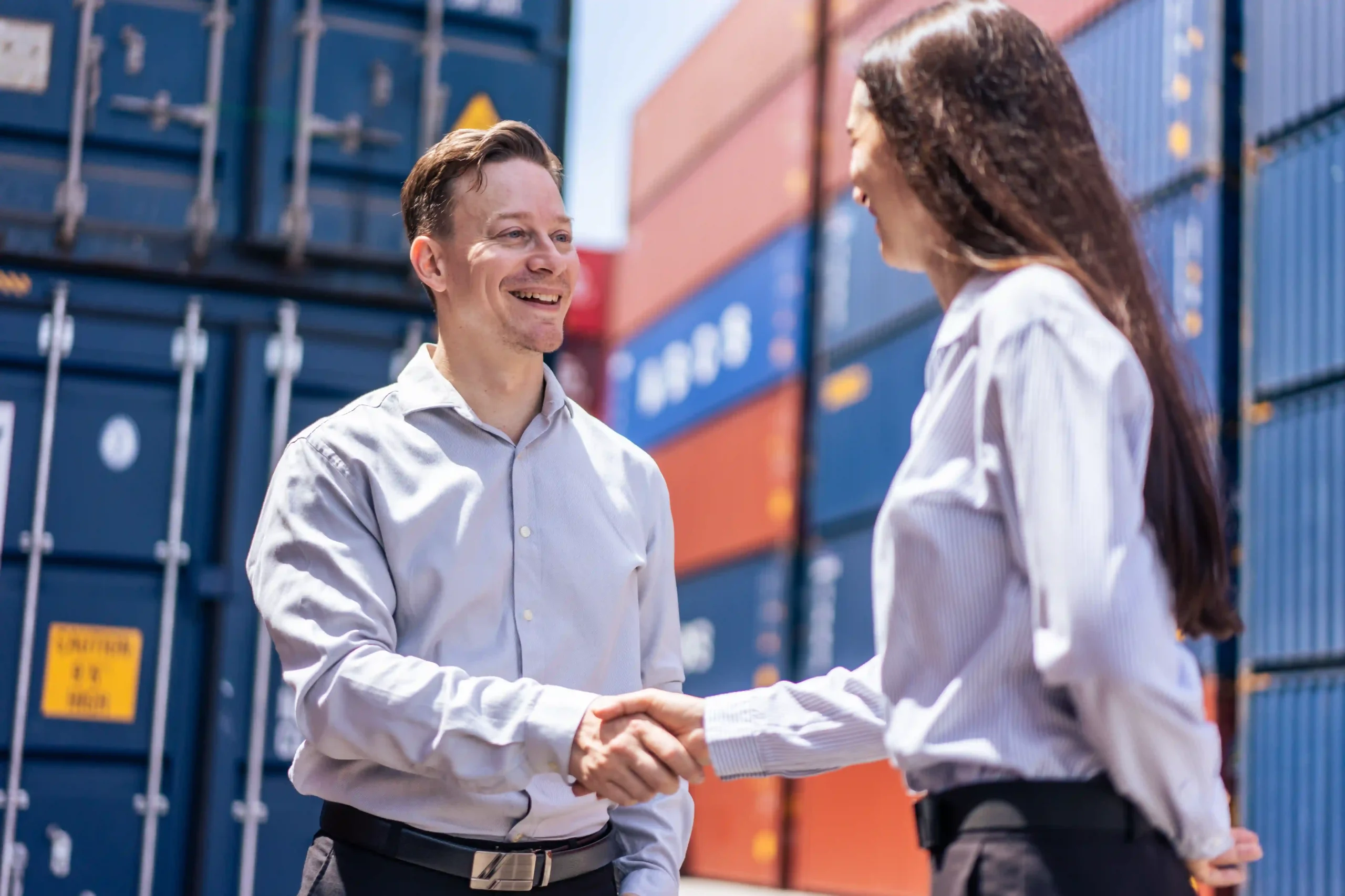 A 3pl logistics manager shakes hands with a client at a shipping yard, standing in front of stacked cargo containers.