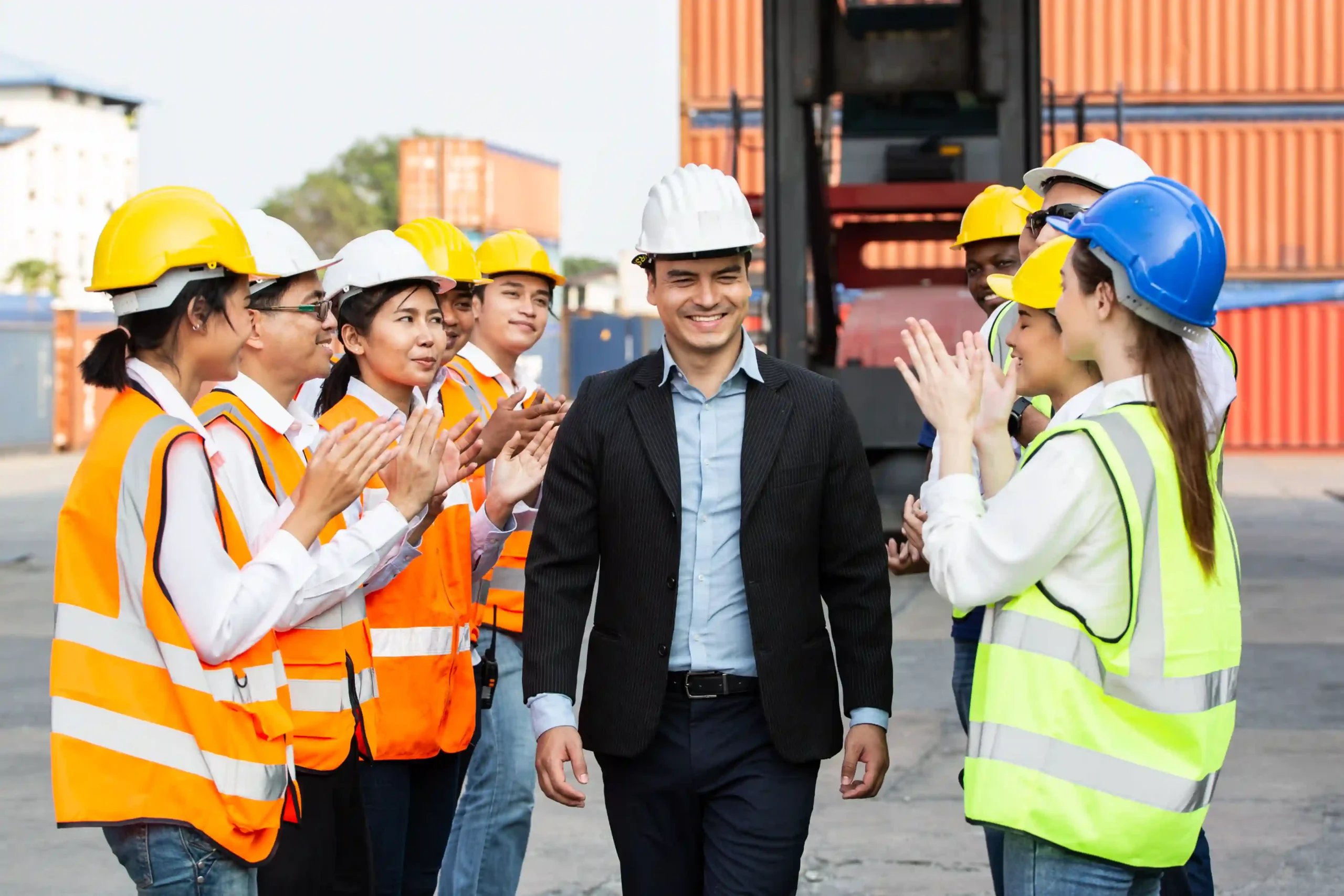 A 3pl logistics manager in a suit walks through a team of warehouse workers clapping and wearing safety gear near freight containers.