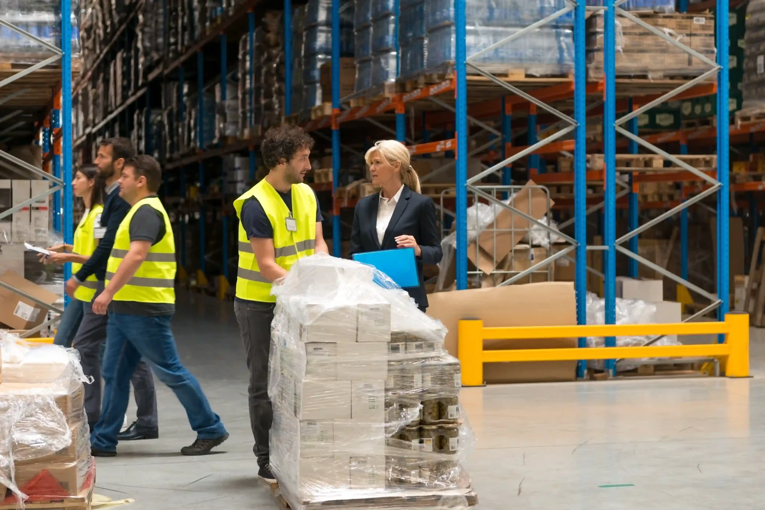 A group of warehouse employees coordinating around a shipment near pallet racks with visible labels, showcasing teamwork and the impact of labeling racks in warehouse for accuracy and efficiency.