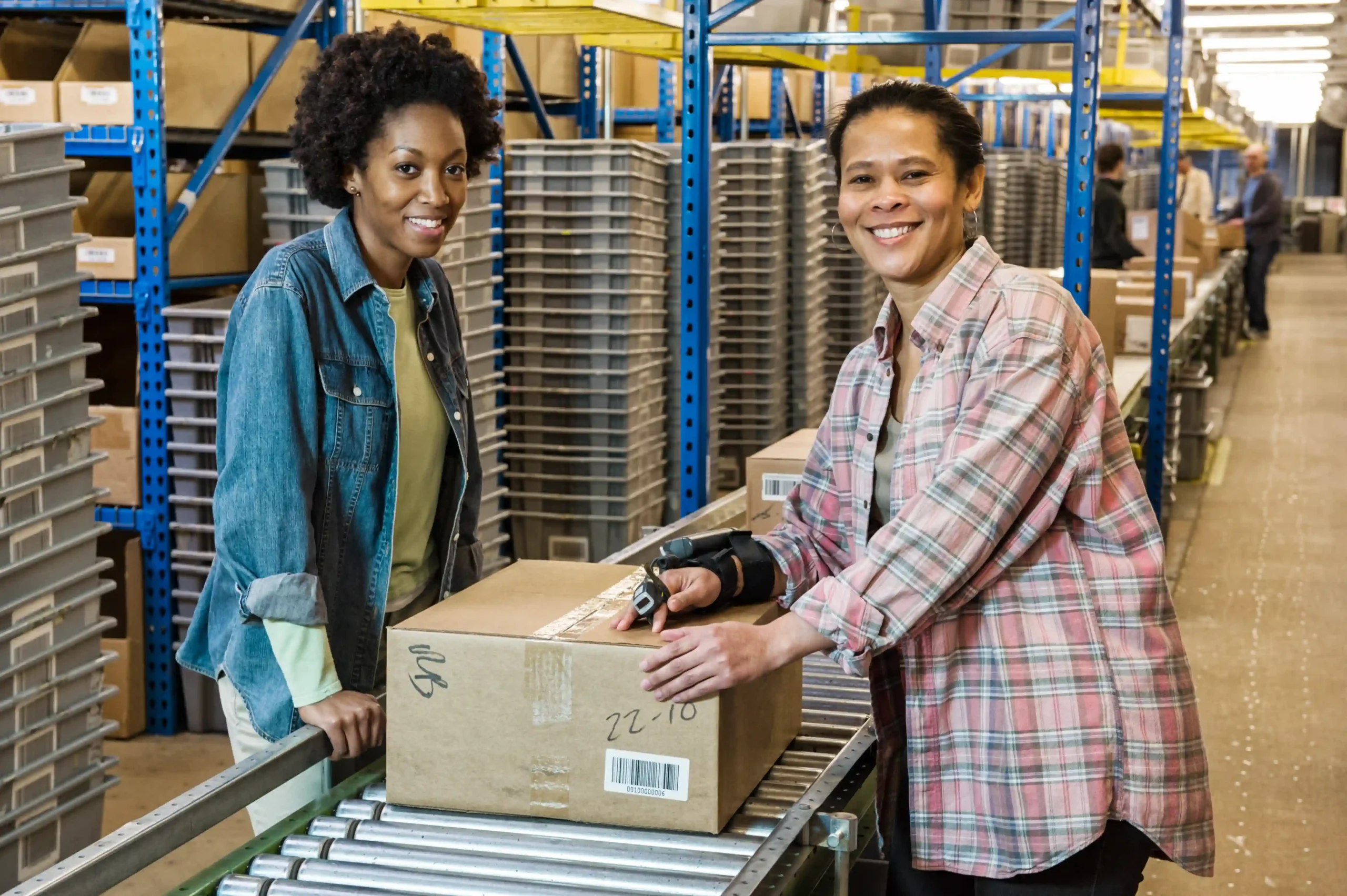 Two smiling warehouse workers standing beside a conveyor belt while preparing a package, illustrating the pick pack meaning in a logistics facility.