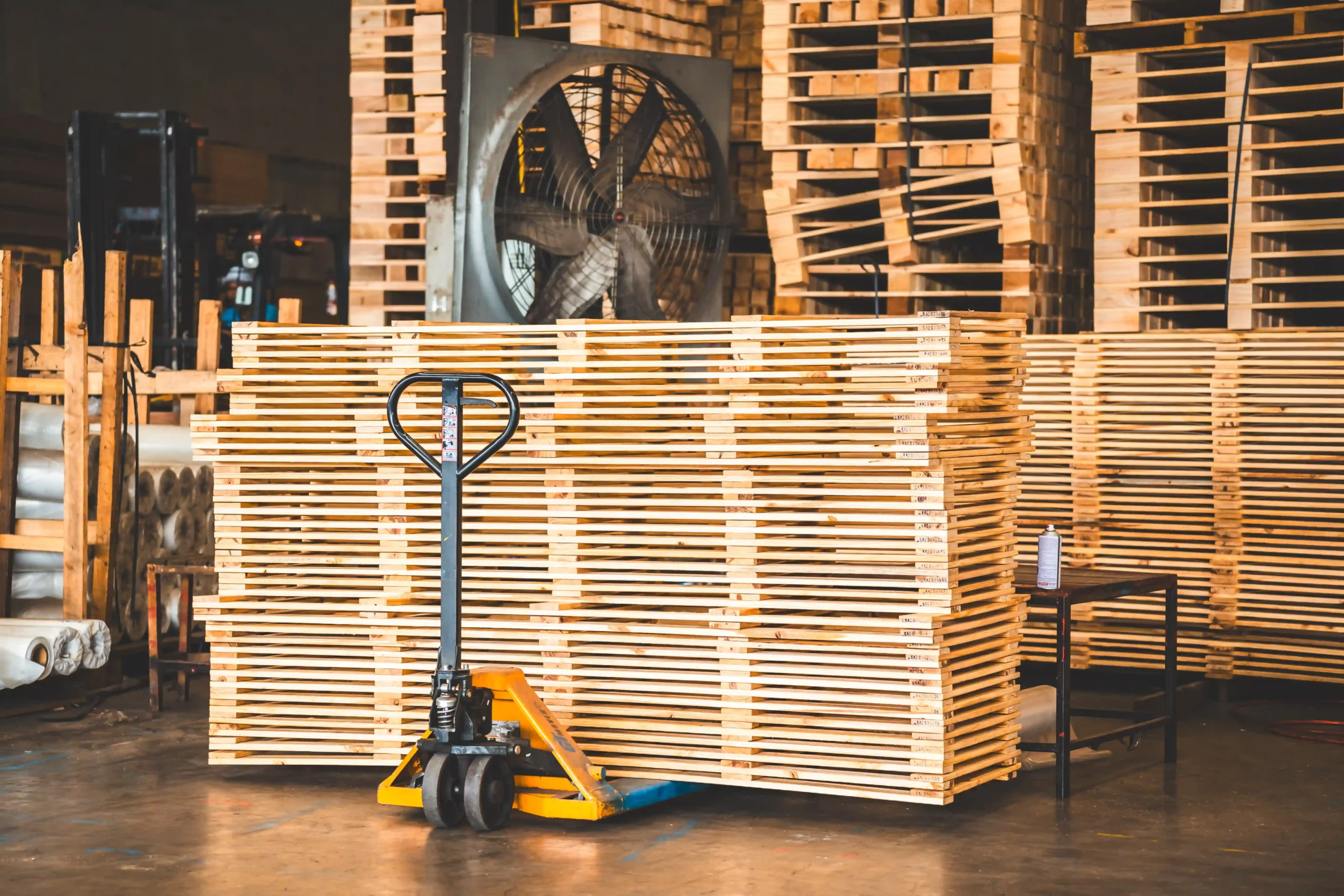 Manual pallet jack beside stacked wood slats in a pallet rack warehouse and supply location.