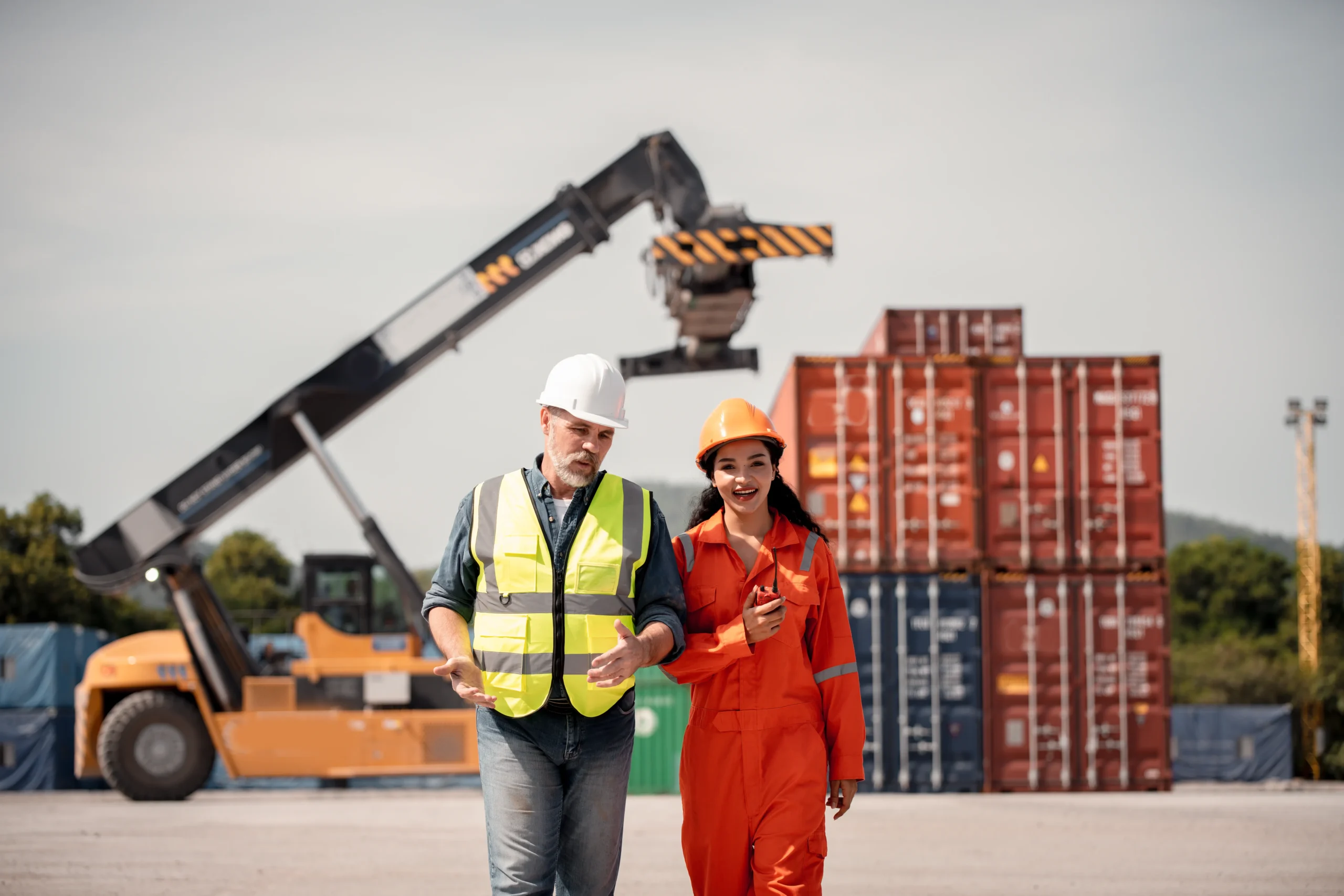 Male and female workers walking through a freight yard with cargo cranes, discussing trade compliance management procedures