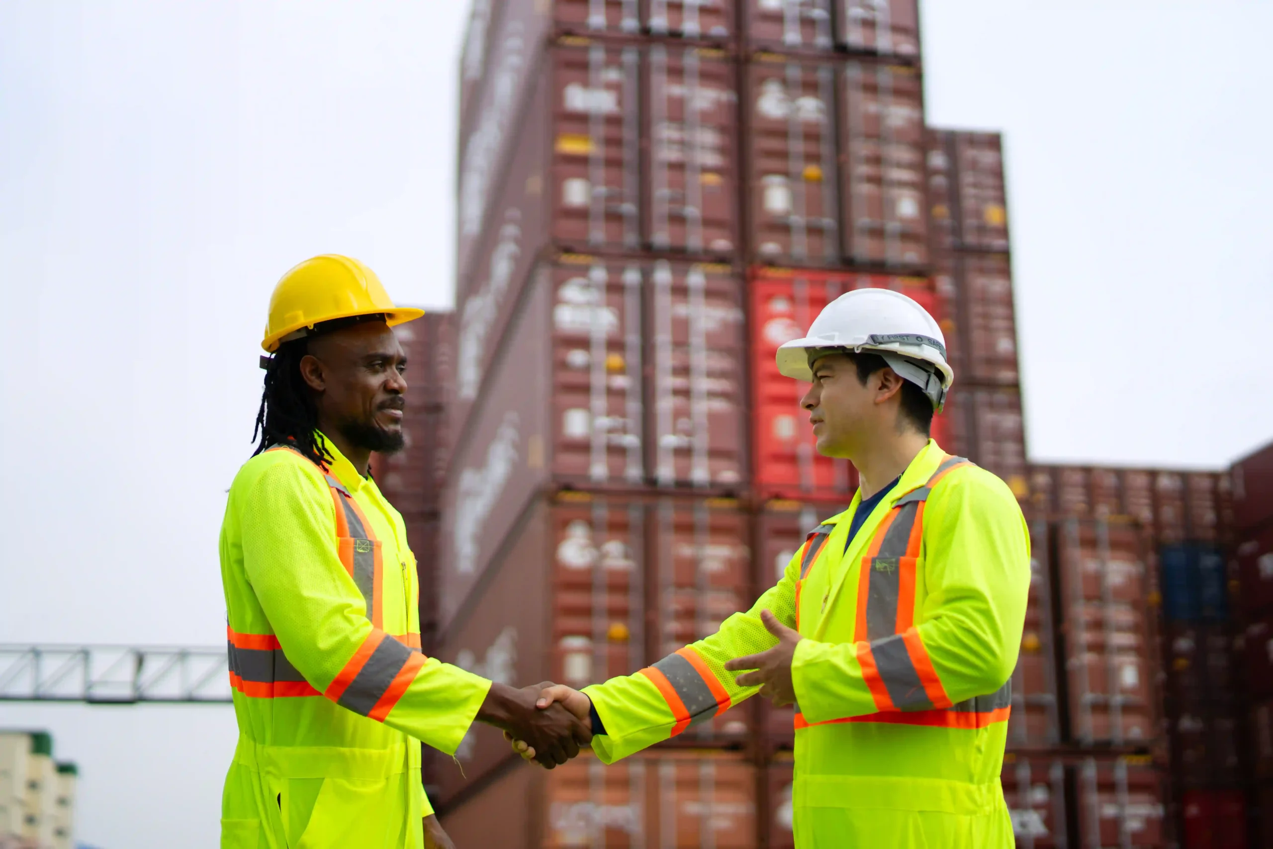 Two logistics workers shake hands in front of stacked shipping containers, symbolizing global trade partnerships under de minimis tariffs.
