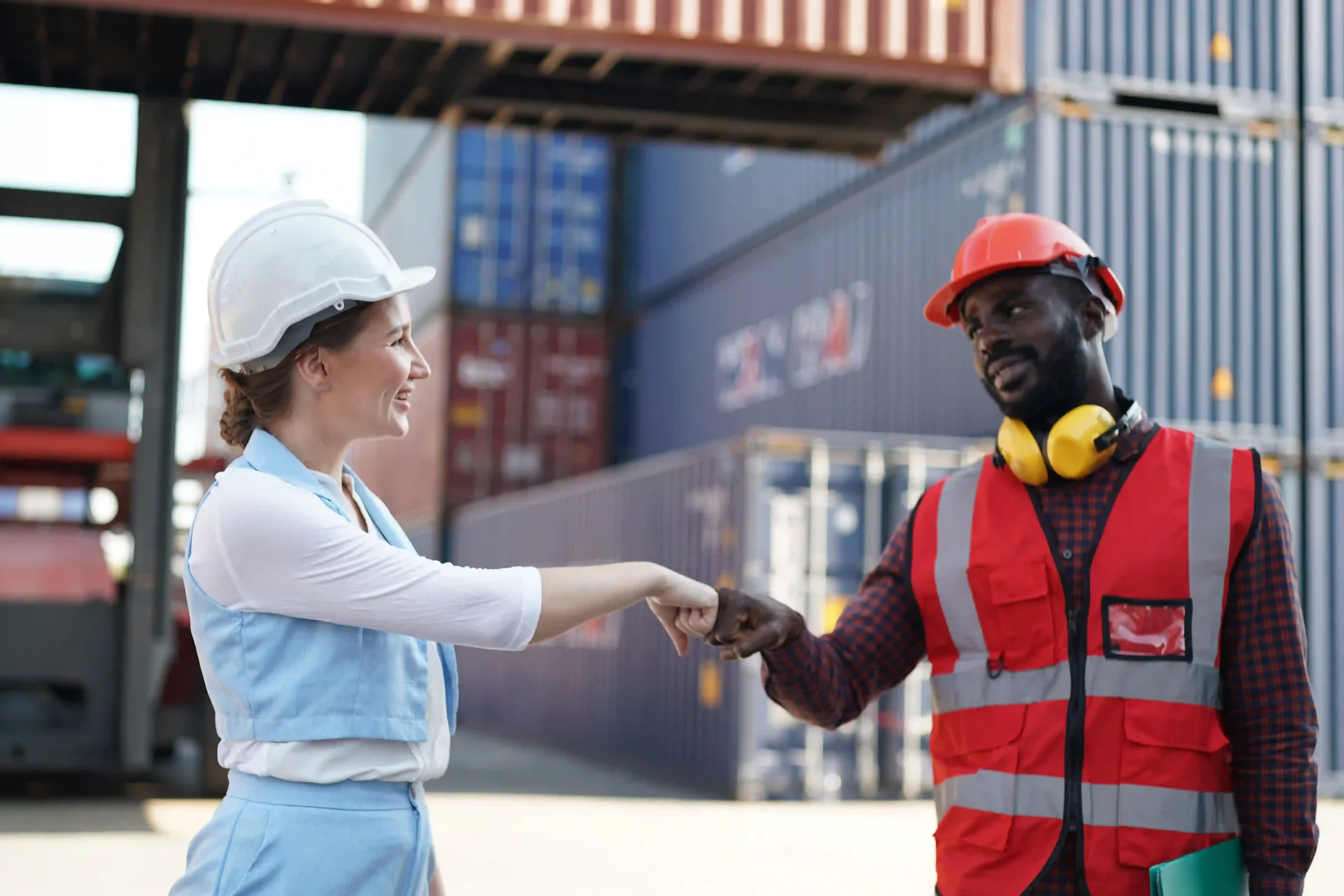 Two logistics workers in safety gear fist bumping in front of shipping containers, symbolizing teamwork under de minimis rule imports.
