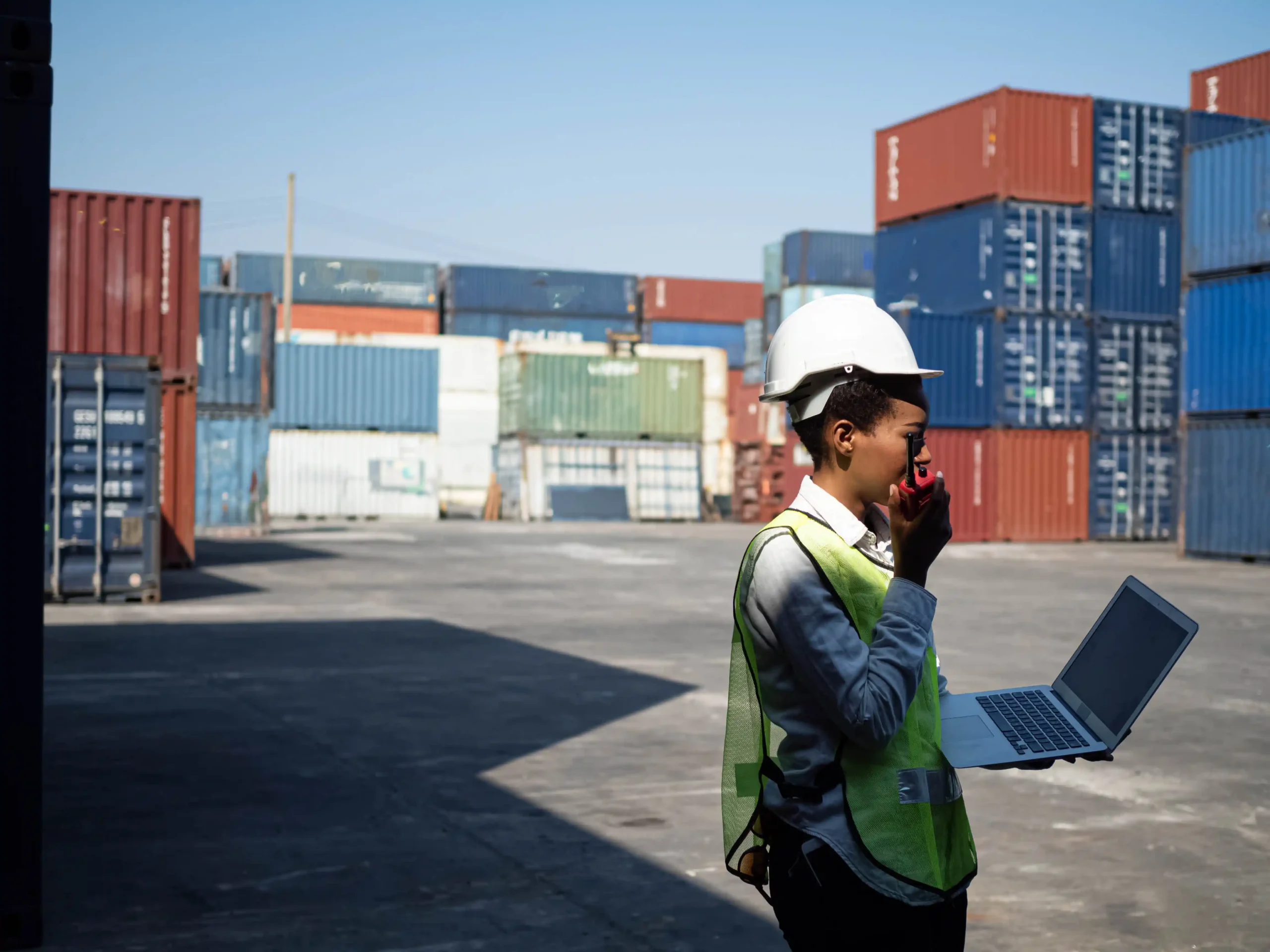 Logistics worker wearing a hardhat and safety vest using a laptop at a container yard, inspecting compliance steps related to the new import duty rules for $800 shipments.