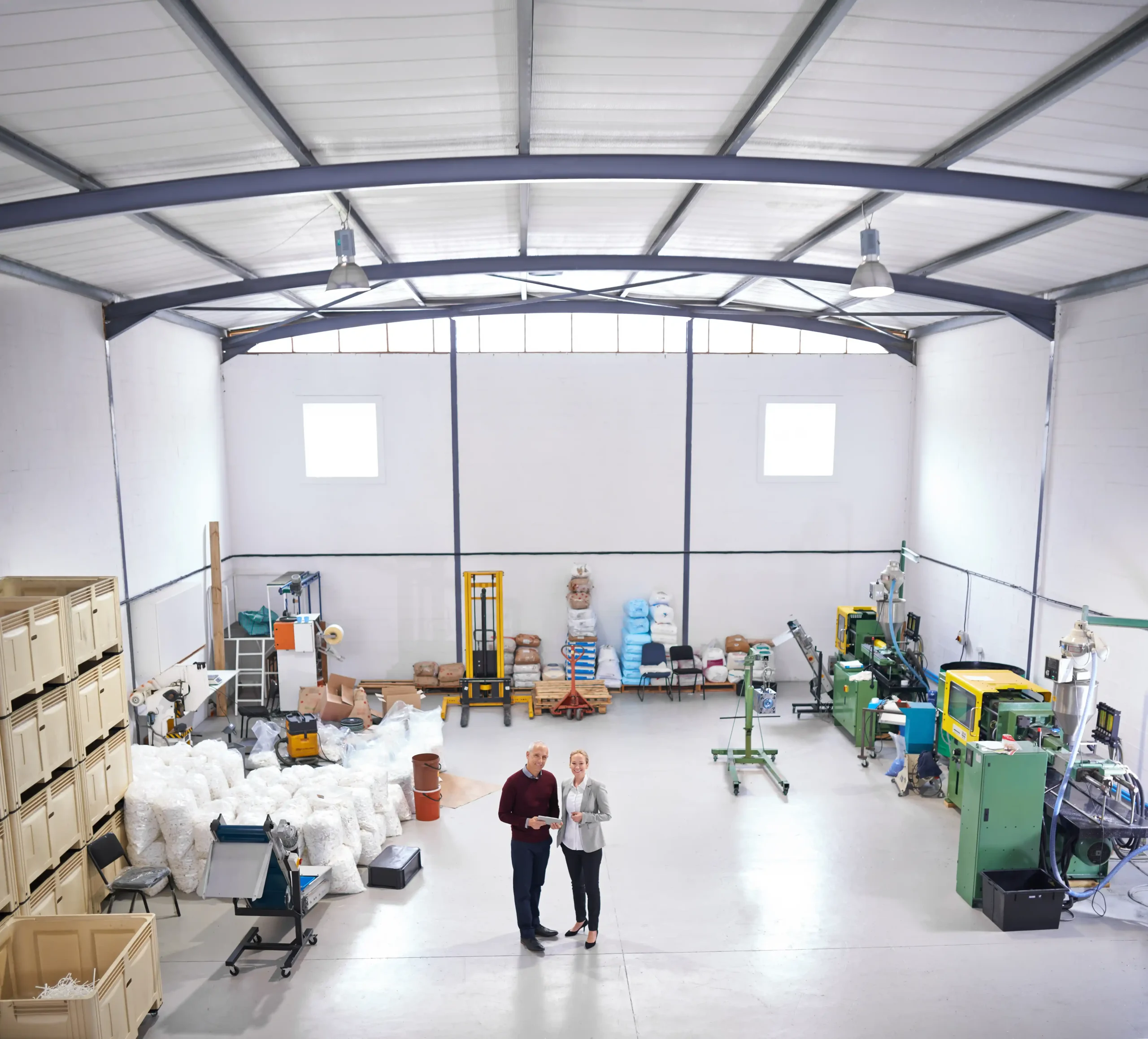 Alt-Text: A small industrial warehouse layout with machinery, stacked materials, and two managers reviewing operations, showcasing efficient little warehouse storage planning.