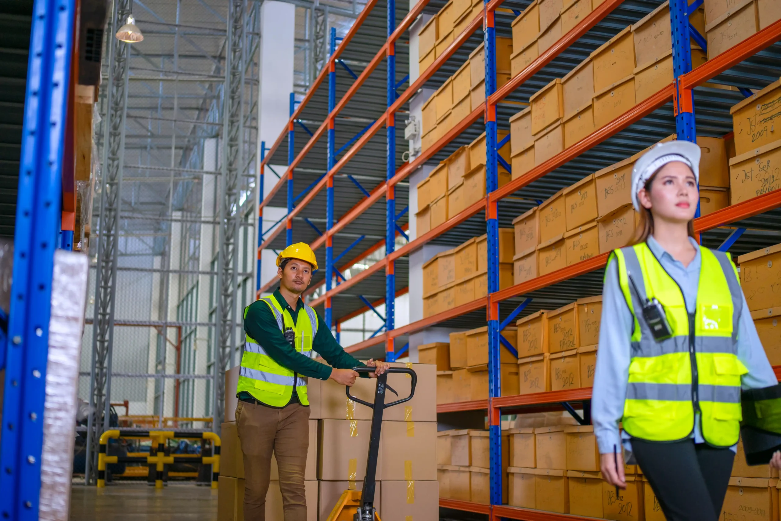 Workers operating equipment in a warehouse storage rack system with tall shelving and organized inventory boxes.