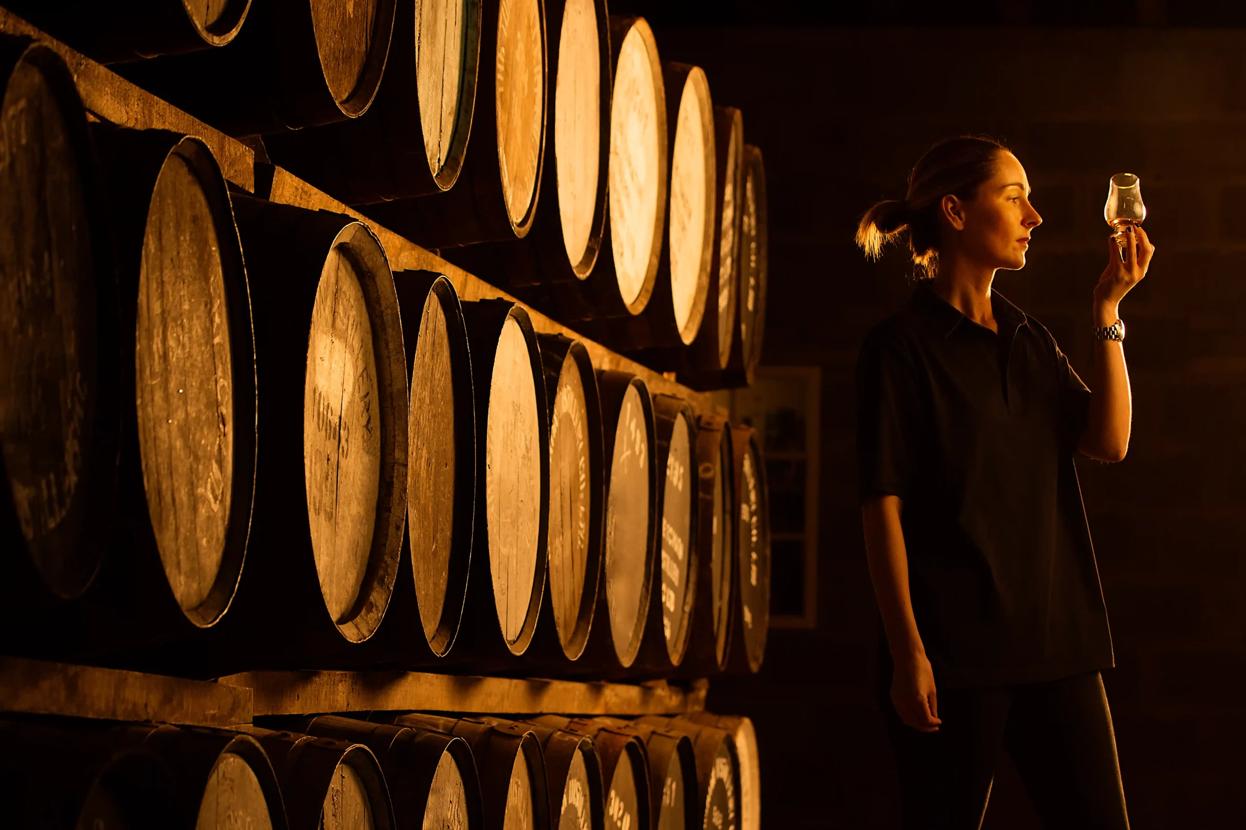 A woman inspecting a glass of wine beside stacked aging barrels, illustrating quality control practices in commercial wine storage settings.