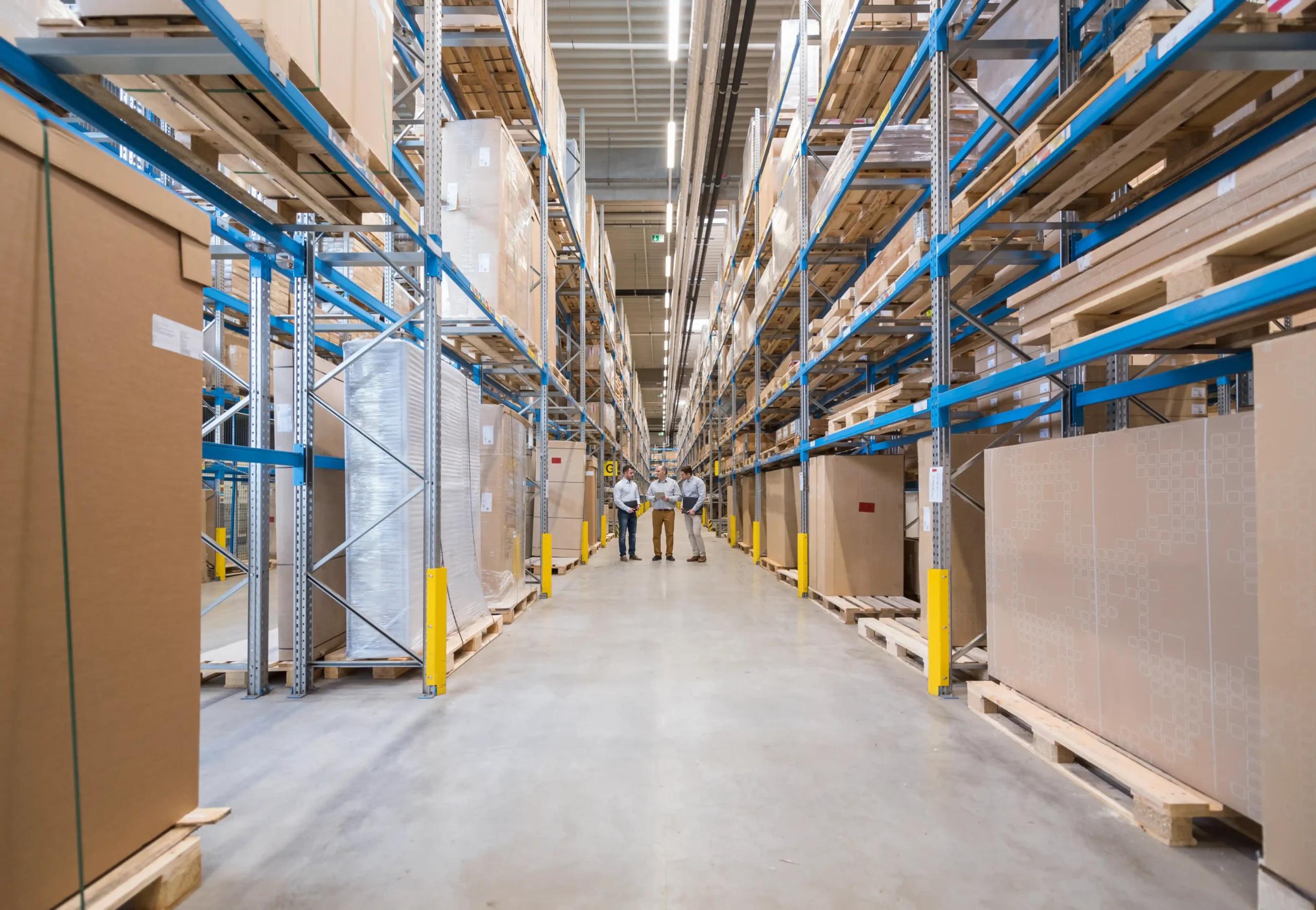 Three supervisors reviewing plans inside a warehouse storage company facility, surrounded by tall shelves and inventory racks.