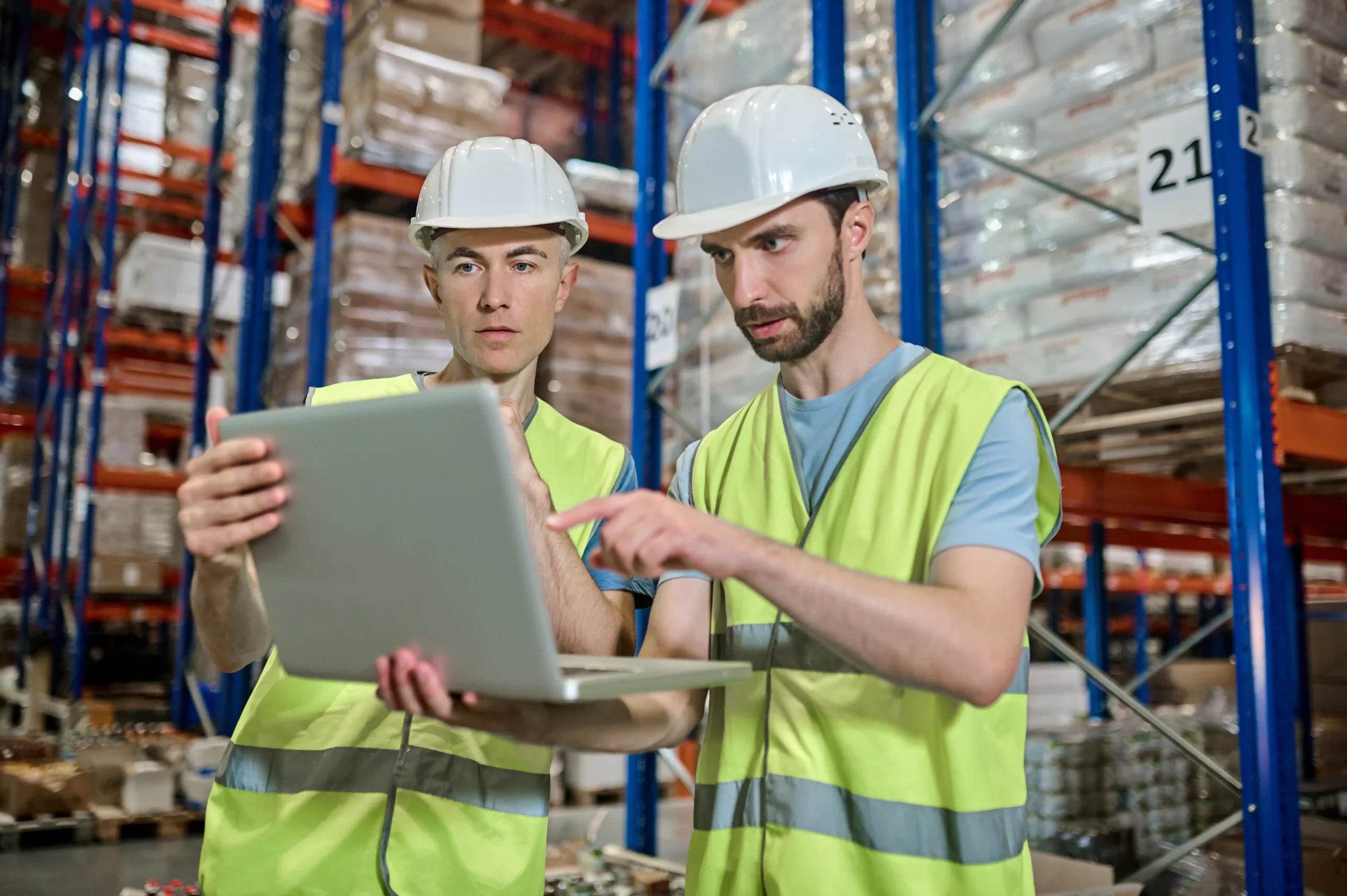 Two warehouse workers in safety gear reviewing inventory data on a laptop, standing in front of warehouse storage racking systems.