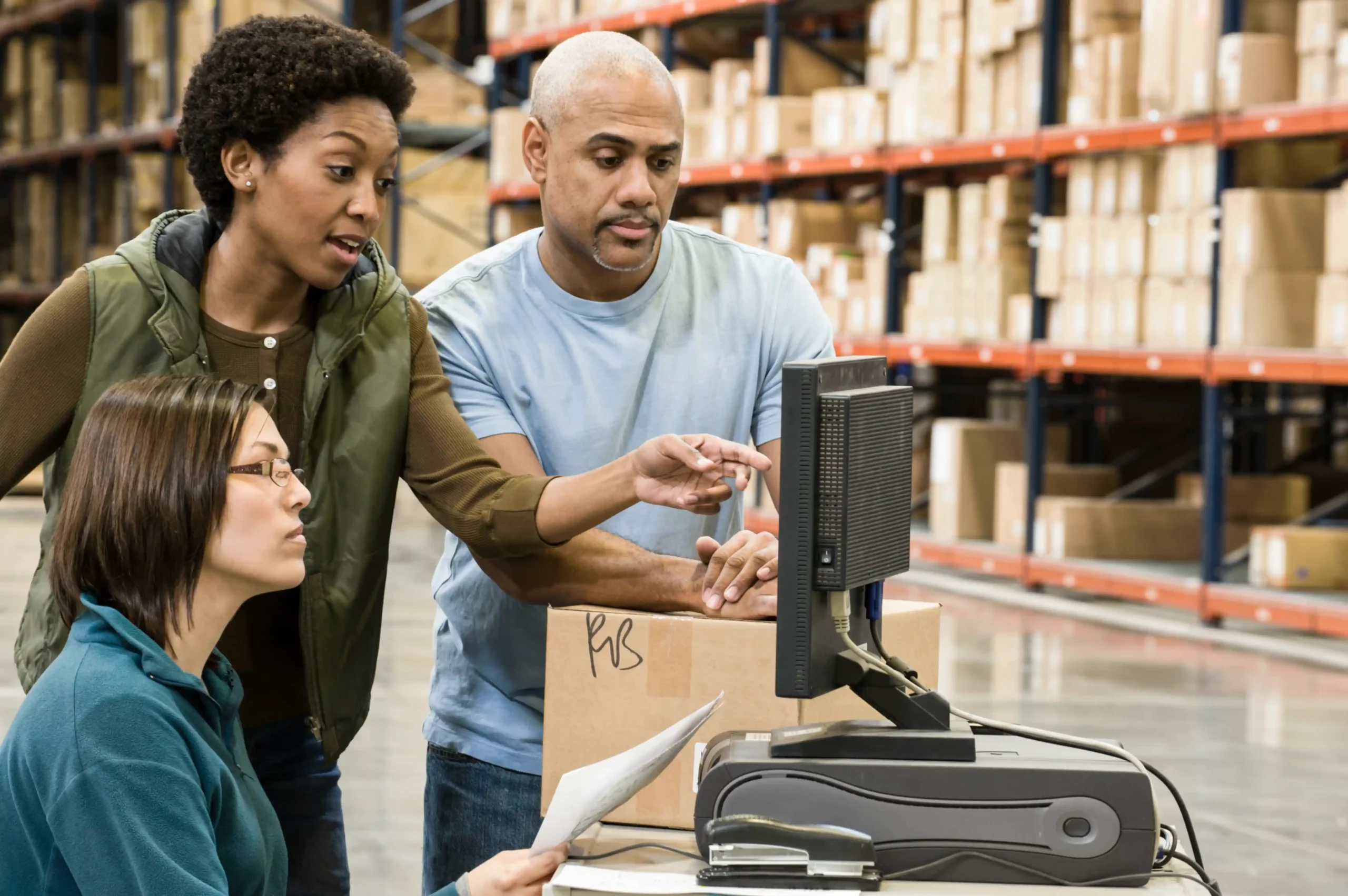Team reviewing inventory details on a computer monitor inside a warehouse with visible warehouse storage racking systems in the background.