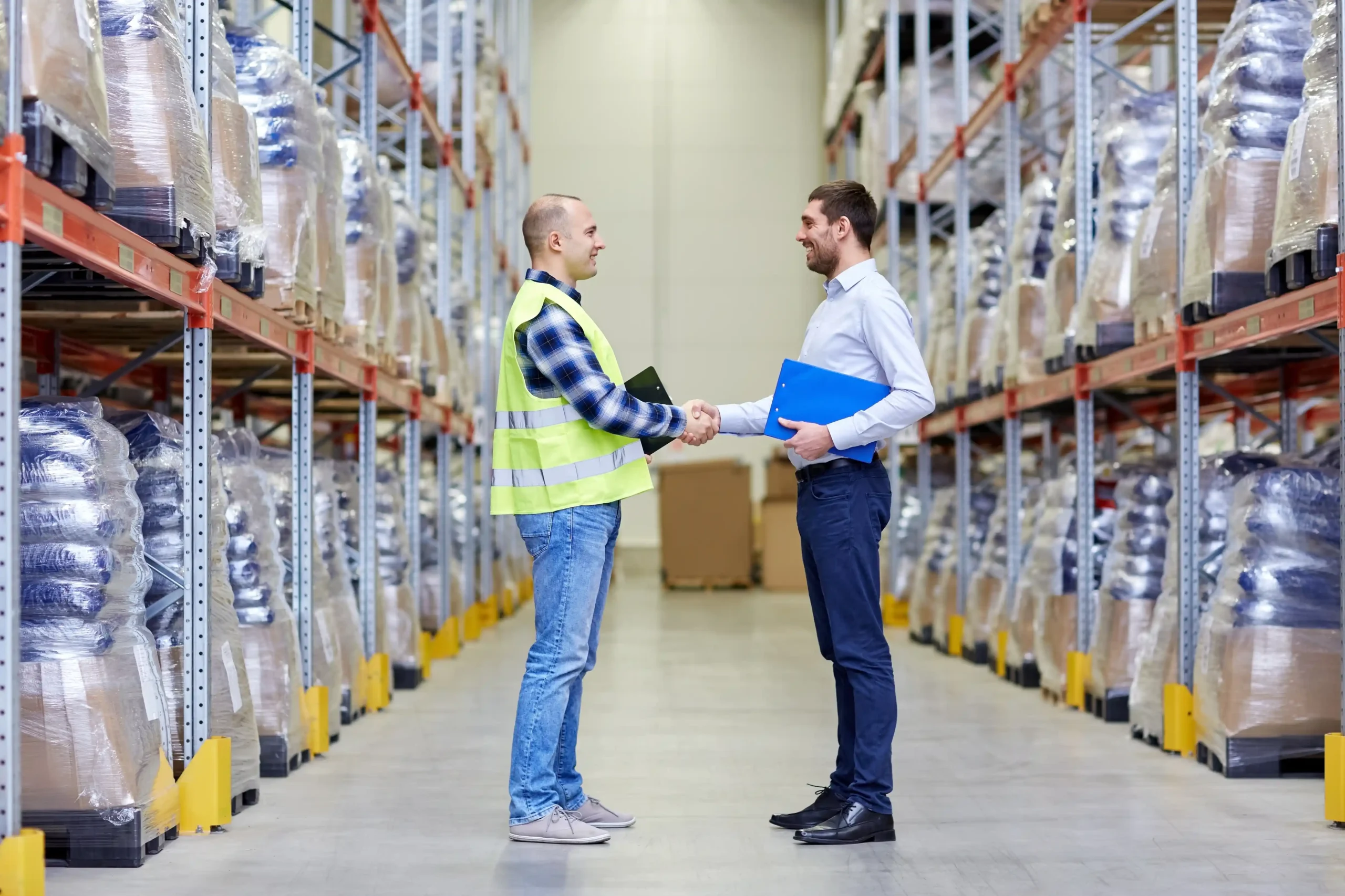 Warehouse employee and business professional shaking hands in an aisle after discussing a warehouse storage agreement with documents in hand.
