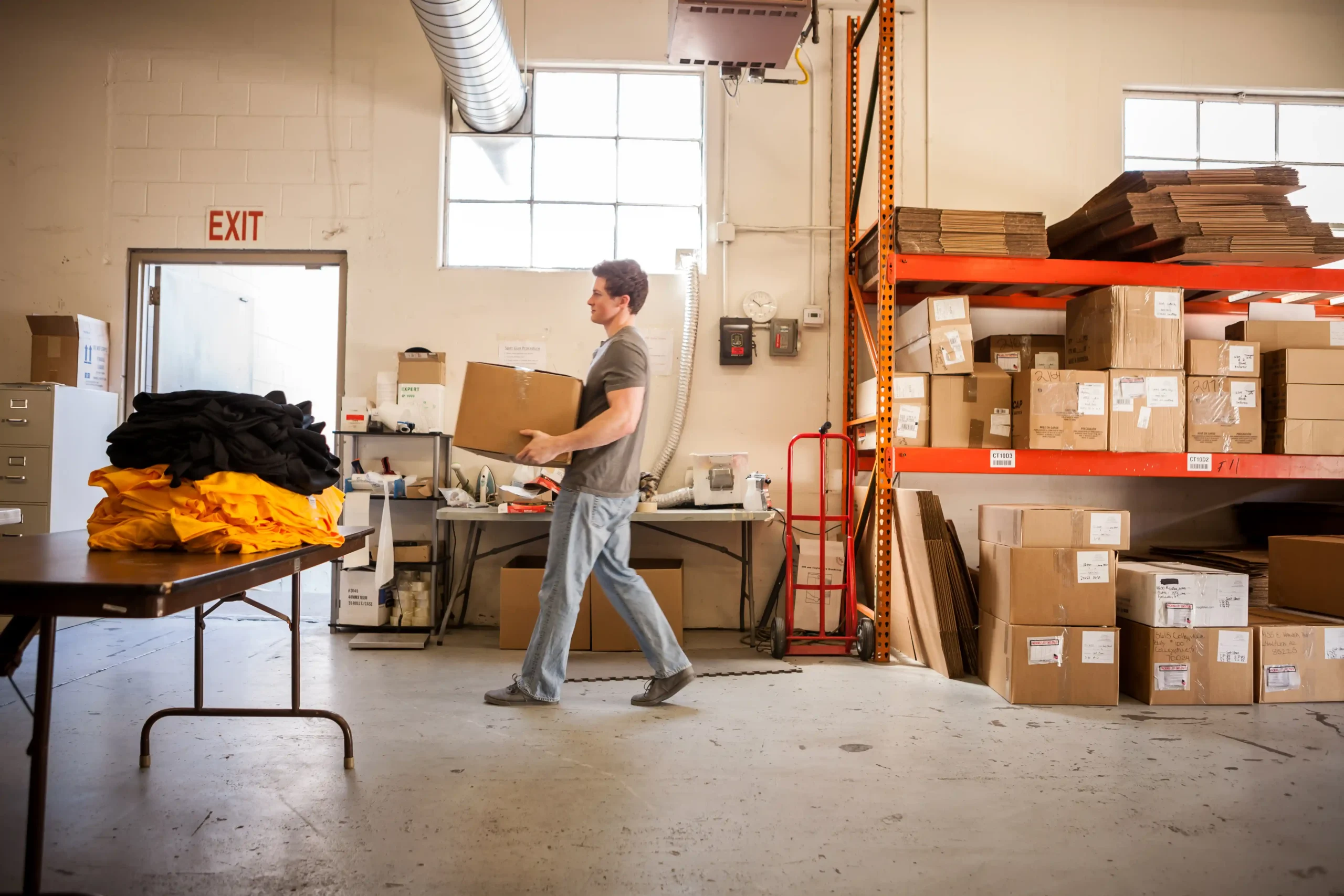 A worker carrying a cardboard box through a warehouse aisle used for clothing warehouse storage and distribution.