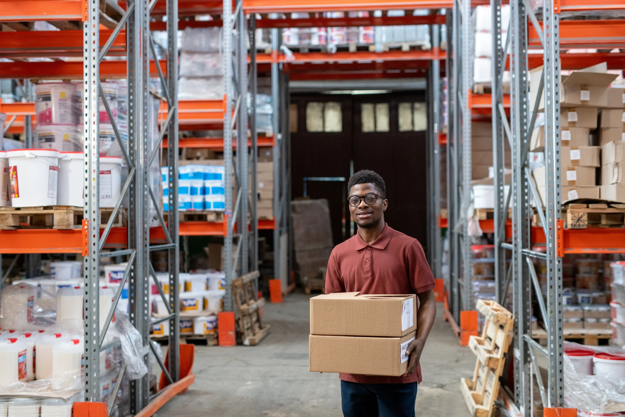 Employee carrying boxes through a warehouse storage zone with organized shelving and labeled inventory sections.