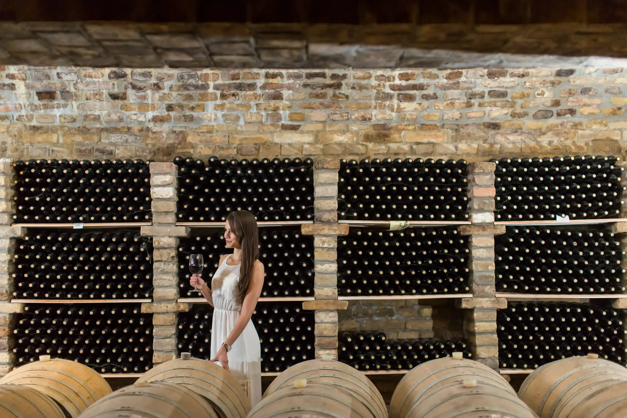 A woman holding a glass of red wine in a historic cellar filled with aging bottles, demonstrating effective commercial wine storage conditions.